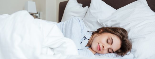 Photo of cheerful young lady dressed in pajama lies in bed at home under a blanket sleeping.
